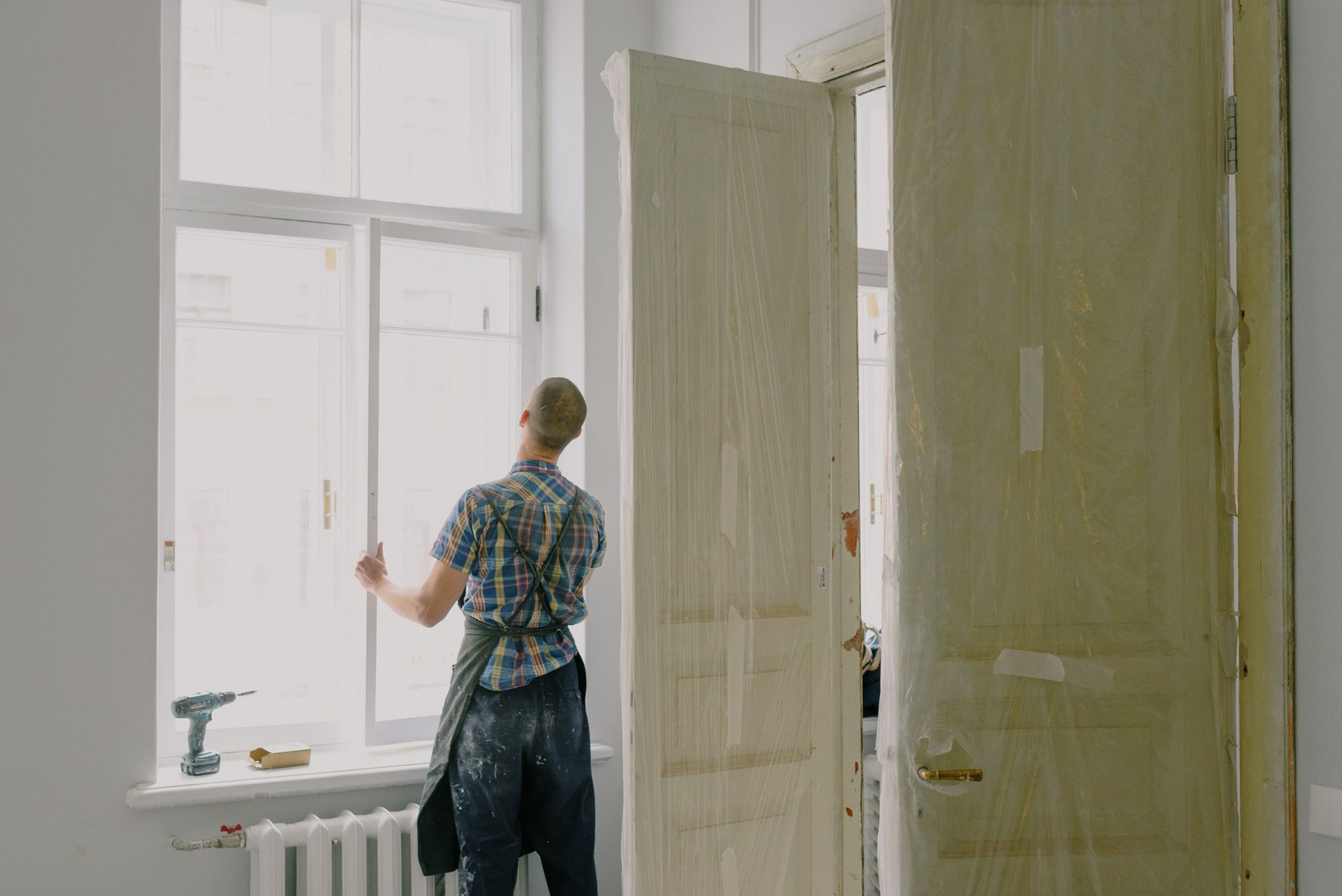 Rückenansicht eines nicht erkennbaren Handwerkers in einer Schürze, der in einer Wohnung ein Fenster montiert – in der Nähe einer Bohrmaschine und von Türen.